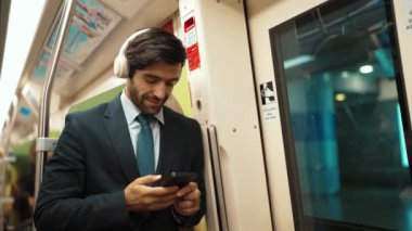 Akıllı yönetici trende dururken kulaklık takar ve müzik dinler. Profesyonel iş adamı, şarkıları dinlerken ve şarkı seçerken bölüme yaslanıyor. İş konsepti. Sevinçli..