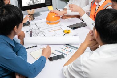 Mimari proje üzerinde birlikte çalışan çeşitli inşaat mühendisi ve müvekkil grupları, inşaat planını gözden geçirip toplantı masasında plan oluşturuyorlar. Tedbirli