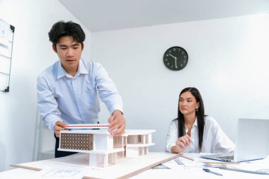 Profesyonel Asyalı erkek mimar ev modelinin uzunluğunu ölçmek için cetvel kullanırken, beyaz tenli genç meslektaşı ev modeliyle toplantı masasındaki verileri analiz etmek için dizüstü bilgisayar kullanıyor. Tertemiz..