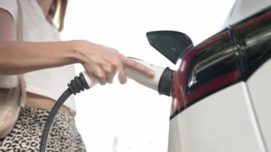 Yavaş çekim güneş ışığı arka plan aydınlatması EV elektrikli arabasıyla gezen kadın yazın yeşil sürdürülebilir şehir bahçesinde şarj oluyor. Elektrikli araç iç organlarından yeşil kentsel sürdürülebilirlik yaşam tarzı