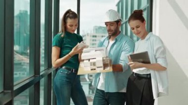 Proje yöneticisi ev modelini elinde tutarken, mimar mühendis takımı da ev tasarımı veya bina inşaatı hakkında fikir alışverişinde bulunuyor. Şehrin penceresinin yanında iç mimar duruyor. İzleme
