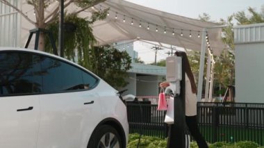 Yavaş çekimde çalışan kadın, otoparktaki EV araba aküsünü alışveriş çantasıyla şarj ediyor. Modern kadın, şehir içi seyahat tarzında çevre dostu elektrikli araçla alışverişe gider. Yararlı