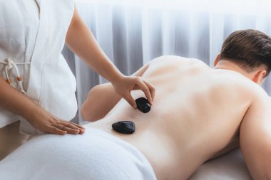 Lüks tatil köyündeki kaplıcada sıcak taş masajı. Gün ışığı dinginliği ortamı, mutlu erkek müşteri bazalt taşı masajının keyfini çıkarıyor. Rahatlatıcı bir sıcaklıkla vücudun üzerinde süzülüyor. Sessiz.