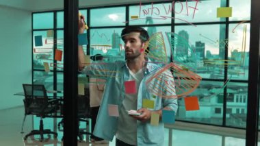 Profesyonel işadamı cam duvara yapışkan notlar yapıştırarak fikir üretmeye çalışırken, aynı zamanda çok kültürlü iş adamları da şehir manzaralı pazarlama fikirlerini tartışıyor. İzleme