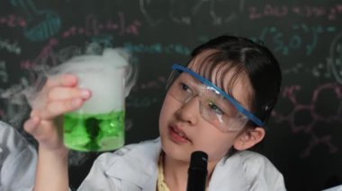 Yeşil kimyasal çözeltiyi elinde tutan genç bir kız ve laboratuvarda biyokimyasal bir teoriyle karatahtanın önünde otururken dikkatlice incele. Deney yapan genç öğrencinin yakın çekimi. Öğretim