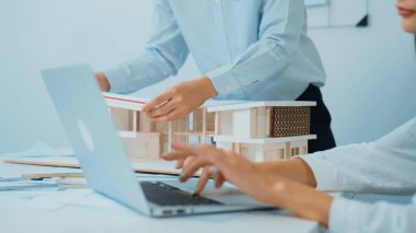 Ev modelinin uzunluğunu ölçmek için cetvel kullanan profesyonel erkek mimarın yakın plan resmi. Bu arada beyaz tenli genç iş arkadaşı ev modeliyle toplantı masasındaki verileri analiz etmek için dizüstü bilgisayar kullanıyor.