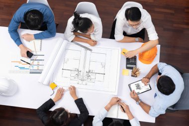 Mimari proje üzerinde birlikte çalışan, inşaat planını gözden geçiren ve toplantı masasında plan oluşturan çeşitli inşaat mühendisi ve müşteri gruplarından oluşan üst düzey bir pankart. Tedbirli