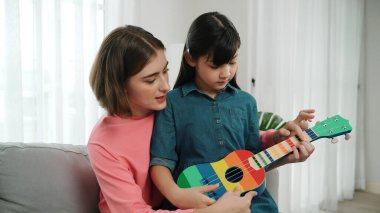Kafkasyalı bir anne evde akustik müzik öğretirken ukulele çalan mutlu kız. Sevimli çocuk enstrüman öğreniyor. Kafkasyalı mutlu anne ve kız birlikte vakit geçiriyorlar. Pedagoji.