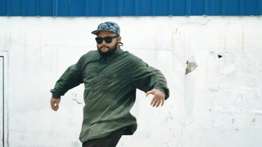Sokak ortasında mavi duvarlı hippi dansçı ayak basamağı. Asyalı break dansçı kollarını açarken sokak dansı yapıyor. Hip hop koreografı enerjik bir dans sergiliyor. Açık hava sporu 2024 hiphop..