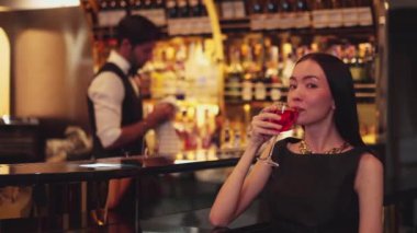 Güzel bir konuk, gece kulübünün mükemmel atmosferinde barmenlerin arka planında çiçeklerle süslenmiş karışık votka Cosmopolitan kırmızı kokteyli ile lüks bir gişede içki içerken kameraya bakıyor. Vinosity.