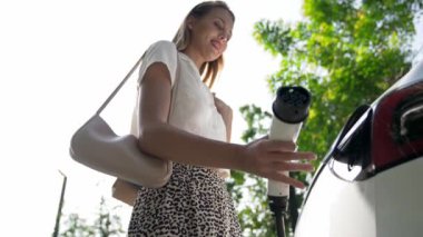 Yavaş çekim güneş ışığı arka plan aydınlatması EV elektrikli arabasıyla gezen kadın yazın yeşil sürdürülebilir şehir bahçesinde şarj oluyor. Elektrikli araç iç organlarından yeşil kentsel sürdürülebilirlik yaşam tarzı