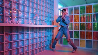 Gece kulübünde kameraya bakarken güneş gözlüklü Asyalı dansçı serbest dans ediyor. Mutlu sokak dansçısı dans ediyor ve renkli parlak neon ışığı ya da yol gösteren ışıkla ayak seslerini gösteriyor. Mutluluk.