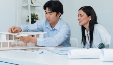 Profesyonel Asyalı erkek mimar ev modelinin uzunluğunu ölçmek için cetvel kullanırken, beyaz tenli genç meslektaşı ev modeliyle toplantı masasındaki verileri analiz etmek için dizüstü bilgisayar kullanıyor. Tertemiz..