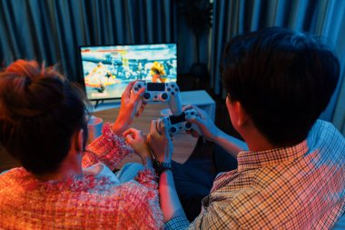 Kontrollü joystickli birkaç oyuncu arka tarafta televizyon ekranında dövüş oyunu oynuyor, modern rahat evinde neon ışıklı renkli oturma odasında koltukta oturuyor. İnfobahn.