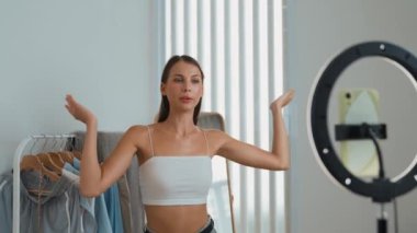 Genç sosyal medyum kadın moda videosu yapıyor. Blogcu kameraya selfie çubuğu ve ışık halkası kullanarak gülümserken, aynı zamanda ikna edici çevrimiçi kıyafetlerin izleyicilere veya takipçilere vlog satmasını sağlıyor. Düzenle