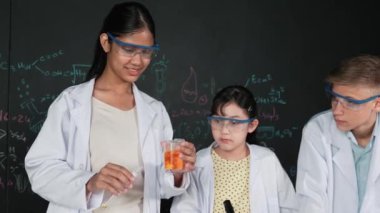 Değişik öğrenciler masada mikroskop ya da tüpü sıvıyla karıştırırken renkli solüsyonları karıştırıyorlar. Kimyasal teorisiyle karatahtanın önünde duran Asyalı kız deney tüpü tutuyor. Öğretim