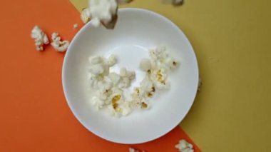 Patlamış mısır, turuncu ve sarı arka planda bir kaseden uçuyor. Beyaz kaseye düşen ve düşen patlamış mısırın yukarıdan görünüşü. Sıradan atıştırmalık konsepti. Poster için tasarım, duvar kağıdı. Pabulum.