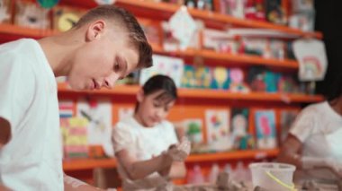 Asyalı lise öğrencisi çömlekçilik atölyesinde çamur ve bir şişe suyla birlikte kil modelliği yapıyor. Bir grup mutlu çocuk çalışıyor ya da bir fincan kil yapıyor. Yaratıcı aktivite konsepti. Öğretim