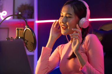 Genç ve güzel Asyalı yayıncının sunucu kanalı, dinleyicilerle sosyal medyada müzik dinliyor, pastel kulaklık takıyor, kapalı alanda neon ışıklı stüdyoda şarkı söylemek için mikrofon kullanıyor. Taktik.