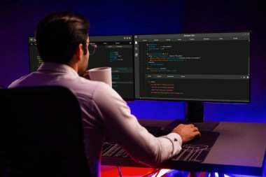Akıllı IT geliştiricisi, bilgisayar ekranında yazılım geliştirme kodlamasında çalışırken kahve fincanı içiyor, program uygulaması güncellemesini neon ofiste sunuyor. Tahmin et..
