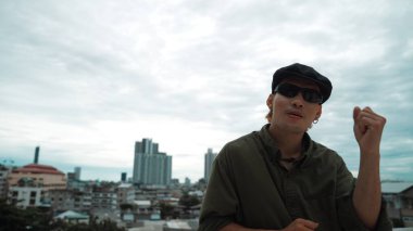 Hipster 'ın yakından çekilmiş görüntüsü kameraya bakarken ellerini çatıya koydu. Hip-hop dansçısı ya da koreograf portresi. Kentle çevrili kameraya bakıyor. Açık hava sporu 2024. Hiphop.