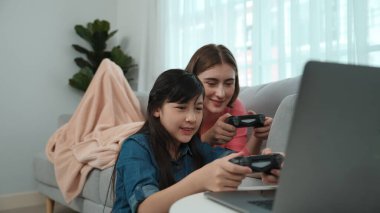 Annem dizüstü bilgisayar kullanarak kanepeye uzanırken, mutlu kız oyun oynuyor ve oturuyor. Beyaz bir ebeveyn ve çekici bir kız, elinde joystick ile birlikte vakit geçirir ve maç kazanmaya odaklanırlar. Pedagoji.