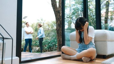 Stresli ve mutsuz genç kız köşeye kıvrılıp kulaklarını kapatarak arka planda ebeveyninin tartışmasını engelliyor. Evde aile içi şiddet ve travmatik çocukluk depresyona dönüşür. Eşzamanlama