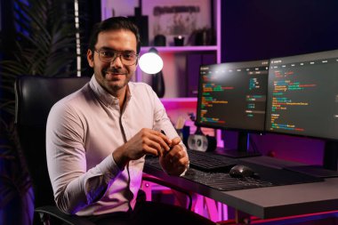 Bilgisayar ekranındaki yazılım geliştirme kodlamasına karşı poz vermek için akıllı bilişim geliştirici görünümlü kamera profilleri, neon ışıklı siber oda ofisinde program güncellemesi internet sitesi verileri sunuyor. Tahmin et..