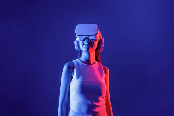 Smart female stand with surrounded by cyberpunk neon light wear VR headset connecting metaverse, futuristic cyberspace community technology. Elegant woman look generated virtual area. Hallucination.
