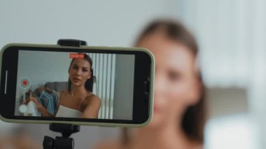 Genç sosyal medyum kadın moda videosu yapıyor. Blogcu kameraya selfie çubuğu ve ışık halkası kullanarak gülümserken, aynı zamanda ikna edici çevrimiçi kıyafetlerin izleyicilere veya takipçilere vlog satmasını sağlıyor. Düzenle