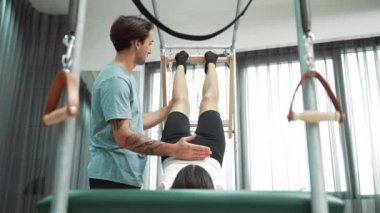 Pilates hocası, Cadillac teçhizatı üzerindeki bacak kaldırma egzersizi sırasında bir müşteriye yardım ediyor. Öğrenci uzanıyor ve temel dayanıklılık ve bacak esnekliğini arttırmak için Cadillac 'ın üzerinde bacak daire çiziyor. Alışkanlık.