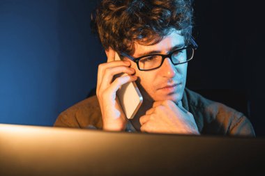 Genç iş adamı akıllı telefondan müşteriyi ya da iş arkadaşını arıyor, neon modern ofisteki yaratıcı sunum surround çalışma masasının bilgisayar ekranına bakıyor. Yeni proje kavramı. Toplayıcı.