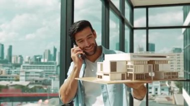 Gülümseyen Kafkasyalı mühendis ev modelini sağa sola çeviriyor ve iş arkadaşına telefon ederken hata noktasını kontrol ediyor. Profesyonel yönetici teftiş, kontrol, mimari modele bakmak. İzleme