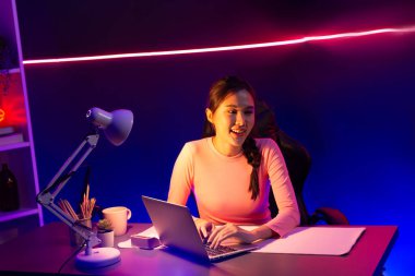 Geceleri sosyal medyada canlı yayında pozitif podcastla konuşan gülümseyen genç, güzel Asyalı etkileyici pembe neon ışıklı modern stüdyoda sunum için dizüstü bilgisayar kullanıyor. Taktik.