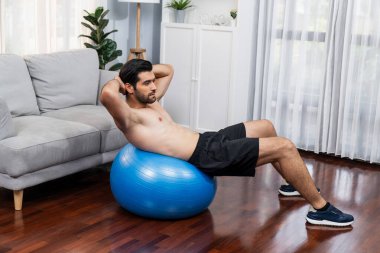 Sporcu ve atletik bir adam evde vücut egzersizi yaparken spor topunun üzerinde oturarak formda fiziki ve sağlıklı spor yaşam tarzına uygun bir yaşam sürüyor. Gaiety ev egzersizi antrenman konsepti.