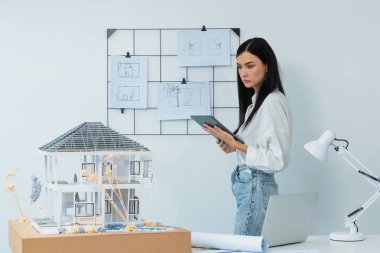 Genç, beyaz, güzel bir mimar, toplantı odasındaki toplantı masasındaki ev modelini, ev modelini ve mimari dökümanları kontrol etmek için tablet kullanmaya odaklanıyor. Tertemiz..