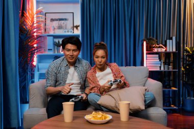 Neşeli, gülümseyen bir çiftin koltuğunda oturup oyun oynuyorlar. Oyun konsolu, hazır aperatifler ve içecekler, modern neon ışıklı, rahat oturma odasında eğlenceli bir rekabet seviyesinde. İnfobahn.