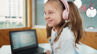 Masaya dizüstü bilgisayarı yerleştirip kameraya el sallayan gülümseyen kız. BTMM sınıfında dizüstü bilgisayar programlama ya da kodlama programı yaparken kulaklık takan bir çocuk gülümsüyor. Etkinlik.