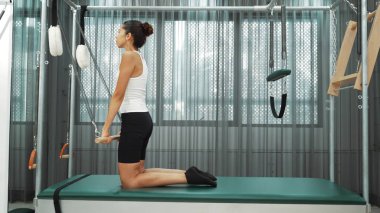 Kadın spor salonunda pilates reformatörüyle çekirdek güçlendirme egzersizi yapıyor. Çekici bayan pilates eğitmeni diz çökmüş vaziyette Cadillac 'ta omuz hareketliliğini arttırmak için bekliyor. Alışkanlık.