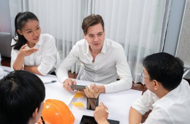 Mimari proje üzerinde birlikte çalışan çeşitli inşaat mühendisi ve müvekkil grupları, inşaat planını gözden geçirip toplantı masasında plan oluşturuyorlar. Tedbirli