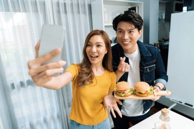 Gülümseyen sunucu şefler akıllı telefonla selfie çekerek sosyal medya kanalından canlı yayında yemek programında çift büyük hamburger yapıyorlar. İnfobahn.