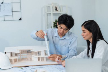 Profesyonel Asyalı erkek mimar ev modelinin uzunluğunu ölçmek için cetvel kullanırken, beyaz tenli genç meslektaşı ev modeliyle toplantı masasındaki verileri analiz etmek için dizüstü bilgisayar kullanıyor. Tertemiz..
