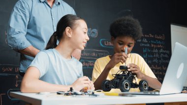 Caucasian teacher looking at laptop screen while explain car model system at blackboard with coding or programing code written. Happy diverse student learning about robotic construction. Edification. clipart