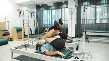 Müşteri, spor salonundaki yenilikçi bir makinede kayışla ters bacak kaldırma hareketi yapıyor. Çekirdek bağlantısını, dengesini ve esnekliğini geliştirmek için ters takla atan iki mutlu antrenör. Alışkanlık.