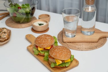 Kare saplı ahşap tahtada çift dev kızarmış hamburger yemeğine odaklanıyorum. Salatayla süslenmiş, ev yapımı çörek, beyaz masadaki fast food konseptinde su içmenin yanı sıra. İnfobahn.