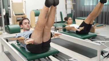 Bir grup müşteri, yenilikçi bir makinede bacak yükseltme işlemi yapıyor. Pilates partneri reformcu pozu veriyor ve egzersiz sırasında dengeyi geliştirmek ve koordinasyonu arttırmak için kayış takıyor. Alışkanlık.