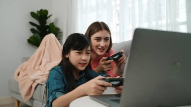 Annem dizüstü bilgisayar kullanarak kanepeye uzanırken, mutlu kız oyun oynuyor ve oturuyor. Beyaz bir ebeveyn ve çekici bir kız, elinde joystick ile birlikte vakit geçirir ve maç kazanmaya odaklanırlar. Pedagoji.