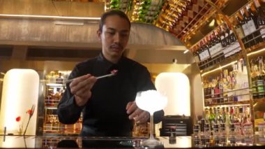 Asyalı barmen karışık kremalı votka bardağı servis ediyor. Önünde buzlu pembe çiçekli şeker, önünde özel içecek, lüks tezgah barında alkolü canlandırıyor. Vinosity.