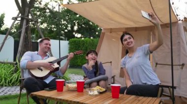 Baba gitar çalarken anne ve kızı bahçede kamp yapıyor. Ebeveynler, genç nesil çapraz kuşak farklılıklarıyla zaman geçirmek için açık hava kamp aktivitelerini kullanıyorlar. Uyumsuzluk.