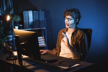 Genç yaratıcı iş adamı, bilgisayar ekranında dinleyicilerle iletişim kuruyor ya da takım üyesiyle görüşüyor. Neon Light Modern Ev Ofisi 'nde kulaklık takıyor. Gece vakti çalışma kavramı. Toplayıcı.